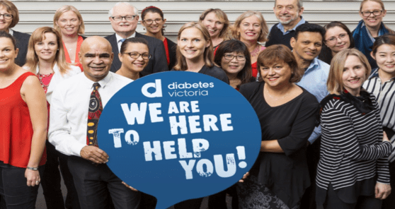 A group of people stand together, smiling, holding a large speech bubble sign that reads We are here to help you! with the Diabetes Victoria logo on it